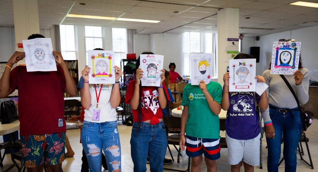 Six kids stand in a classroom with a self-portrait held up over their faces.