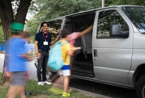 A young man stands by a gray van while students board the vehicle.