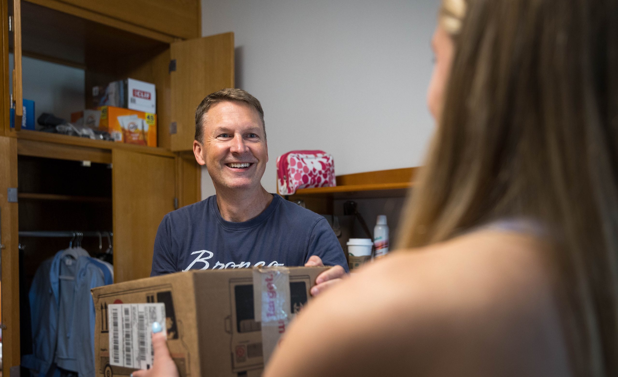 A dad looks on at his daughter, whose back is to the camera, and smiles to her as he holds a box on move-in day in her dorm. 