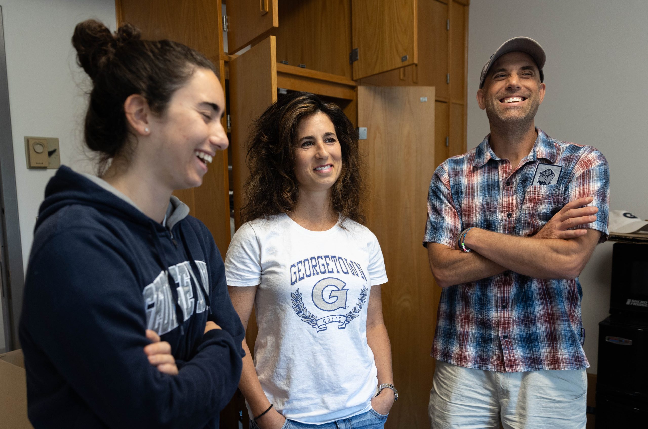 Two parents and their daughter stand together in a dorm room laughing. The daughter is wearing a navy blue Georgetown sweatshirt and the mom a white T-shirt with Georgetown's logo on it.
