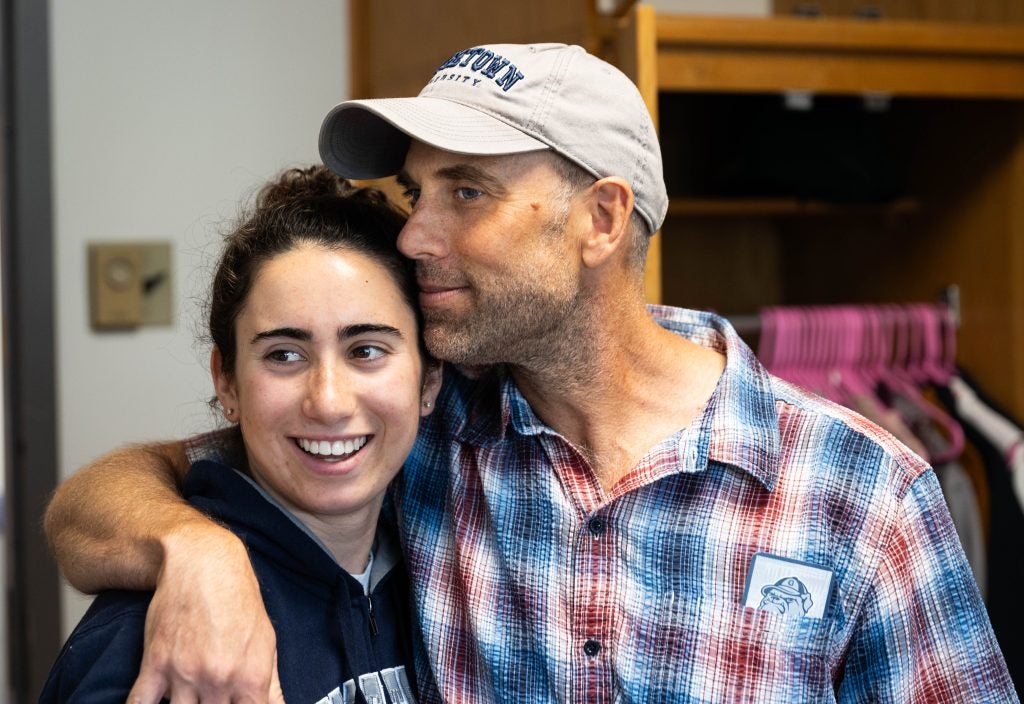 A father wraps his arm around his daughter and leans his head against hers. His daughter smiles off-camera.