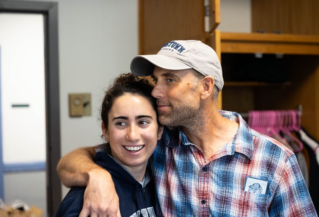 A father wraps his arm around his daughter and leans his head against hers. His daughter smiles off-camera.