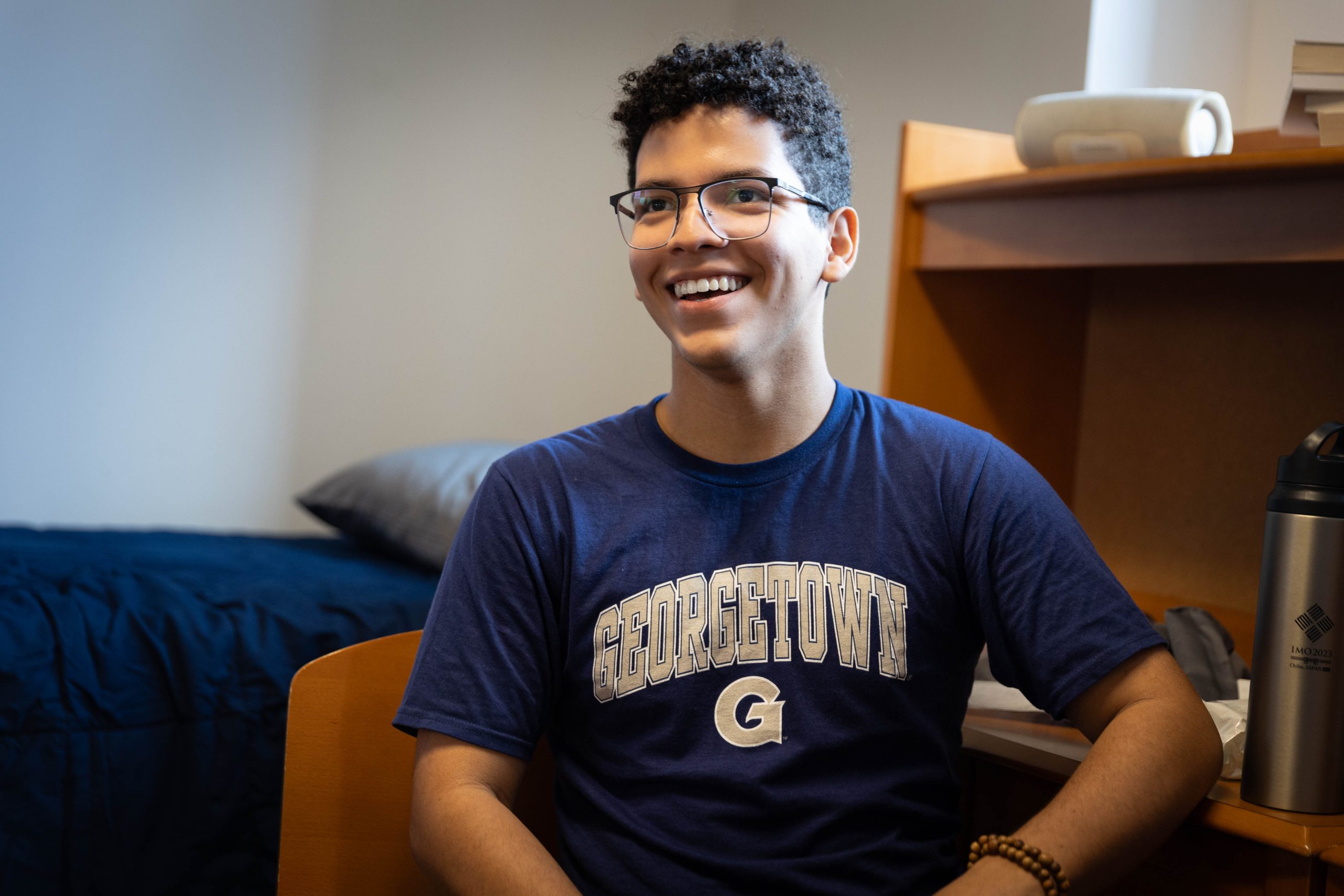 A male college student wearing glasses and a navy blue Georgetown T-shirt smiles off-camera in his dorm room on the first day of move-in.