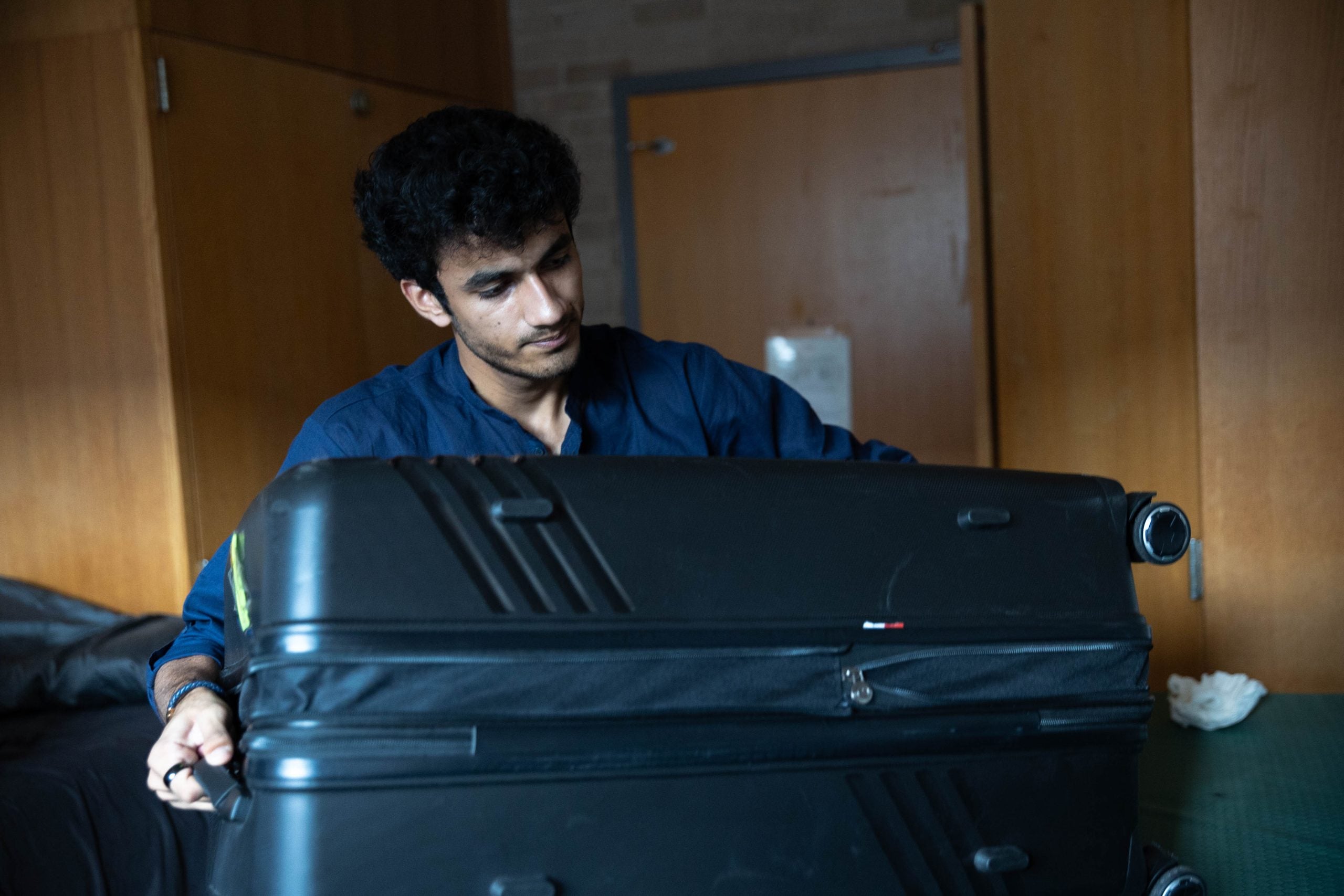 A male college student lugs a large suitcase onto his bed in his dorm room.