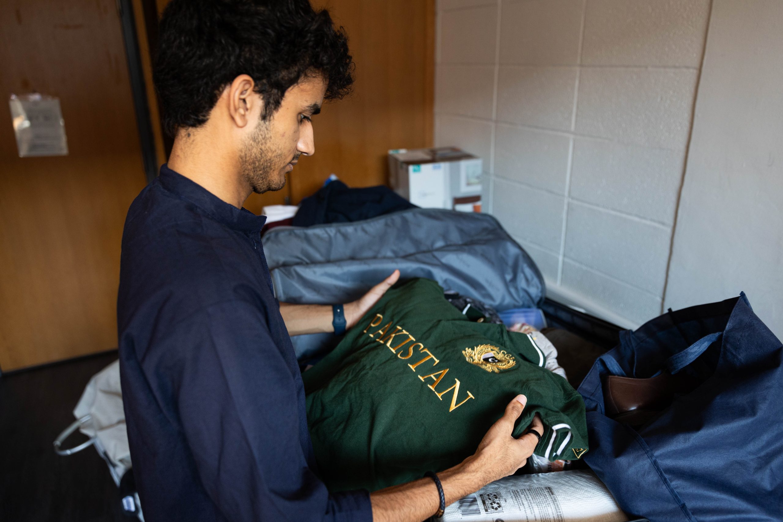 A male college student unpacks a green sweatshirt that says "Pakistan" across the front.