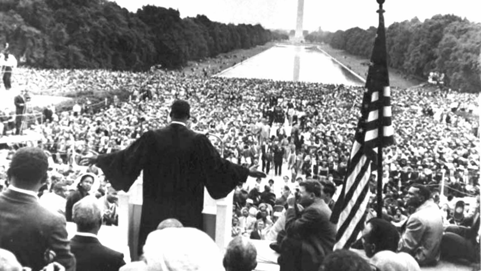 Martin Luther King Jr. addresses crowd of 250,000 from the steps of the Lincoln Memorial. King has arms outstretched and is wearing a black robe with an American flag to his right. Photo is taken from behind with reflecting pool and Washington monument visible in the distance.