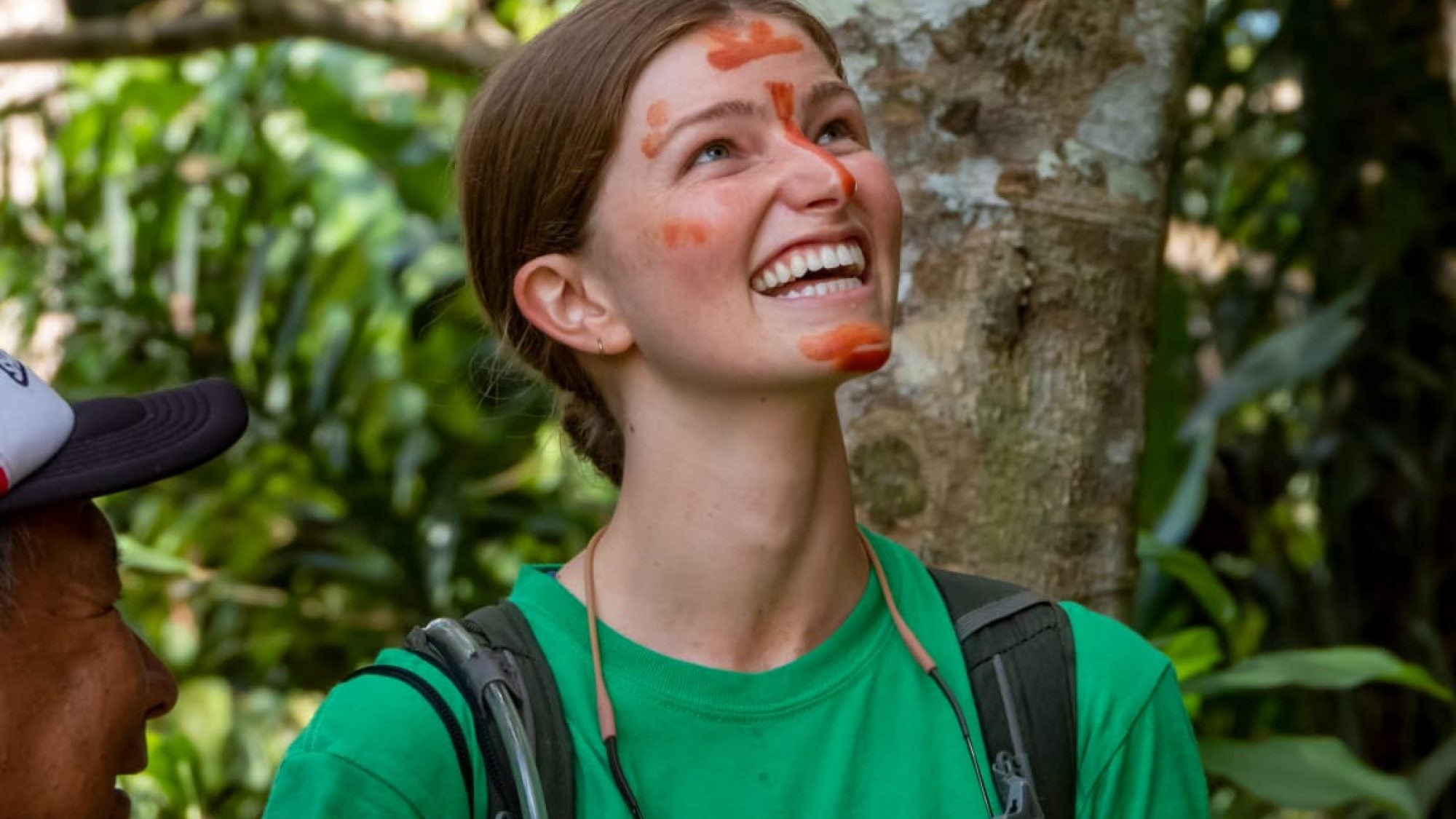 Woman with red face paint in a green shirt looking up in a rainforest