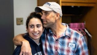 A father wraps his arm around his daughter and leans his head against hers. His daughter smiles off-camera.