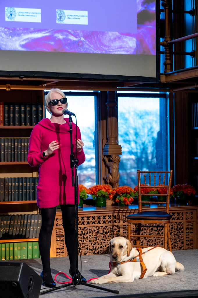 Marissa Nissley (B‘24), a student who is blind, performs on stage with her service dog Smalls at an event on Georgetown's campus celebrating the disability community and the arts. Nissley wears a bright pink dress, black tights and black sunglasses. 