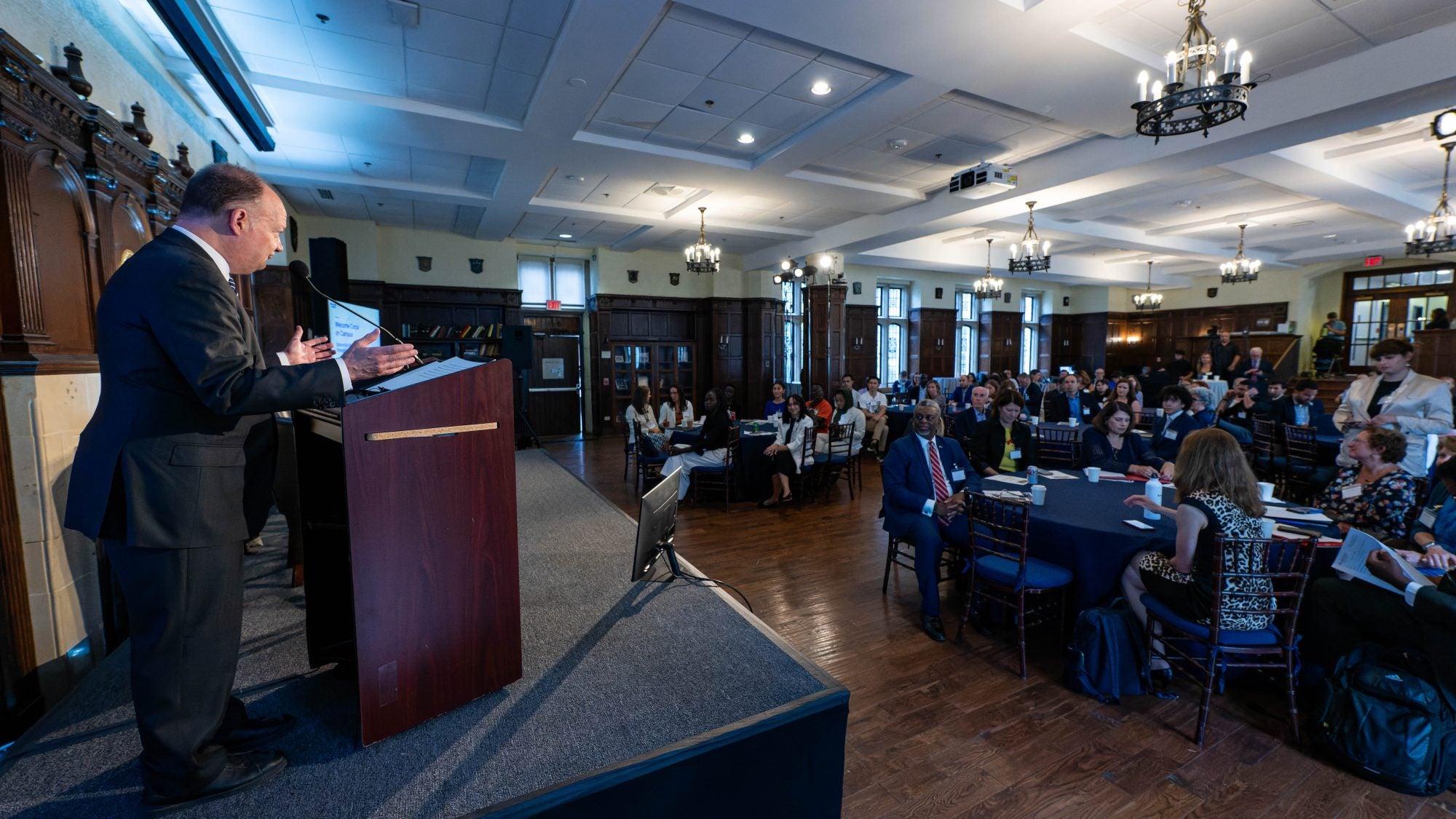 President John J. DeGioia stands behind a podium on a stage in a formal dining room to a crowd of seated attendees.