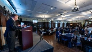 President John J. DeGioia stands behind a podium on a stage in a formal dining room to a crowd of seated attendees.