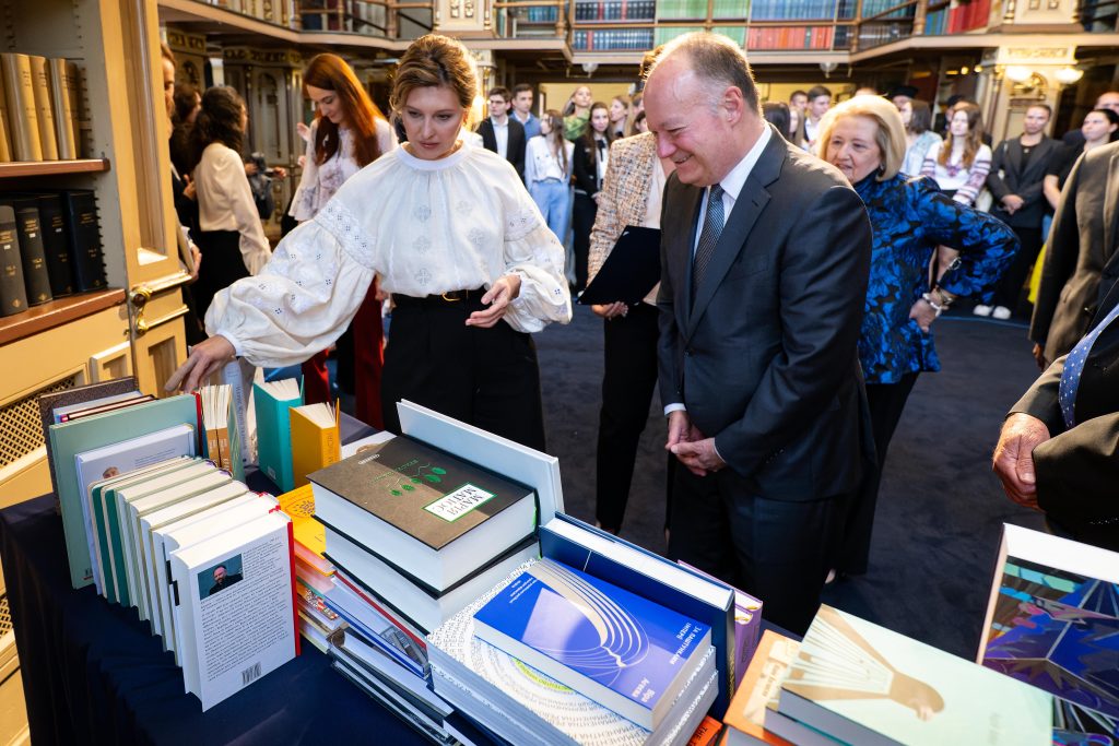 Ukrainian First Lady Olena Zelenska points to a table a books she gifted to Georgetown university alongside President John J. DeGioia. She is wearing a white flowy blouse and the president wears a suit and tie. They are standing with a group of Ukrainian students in a Georgetown library.