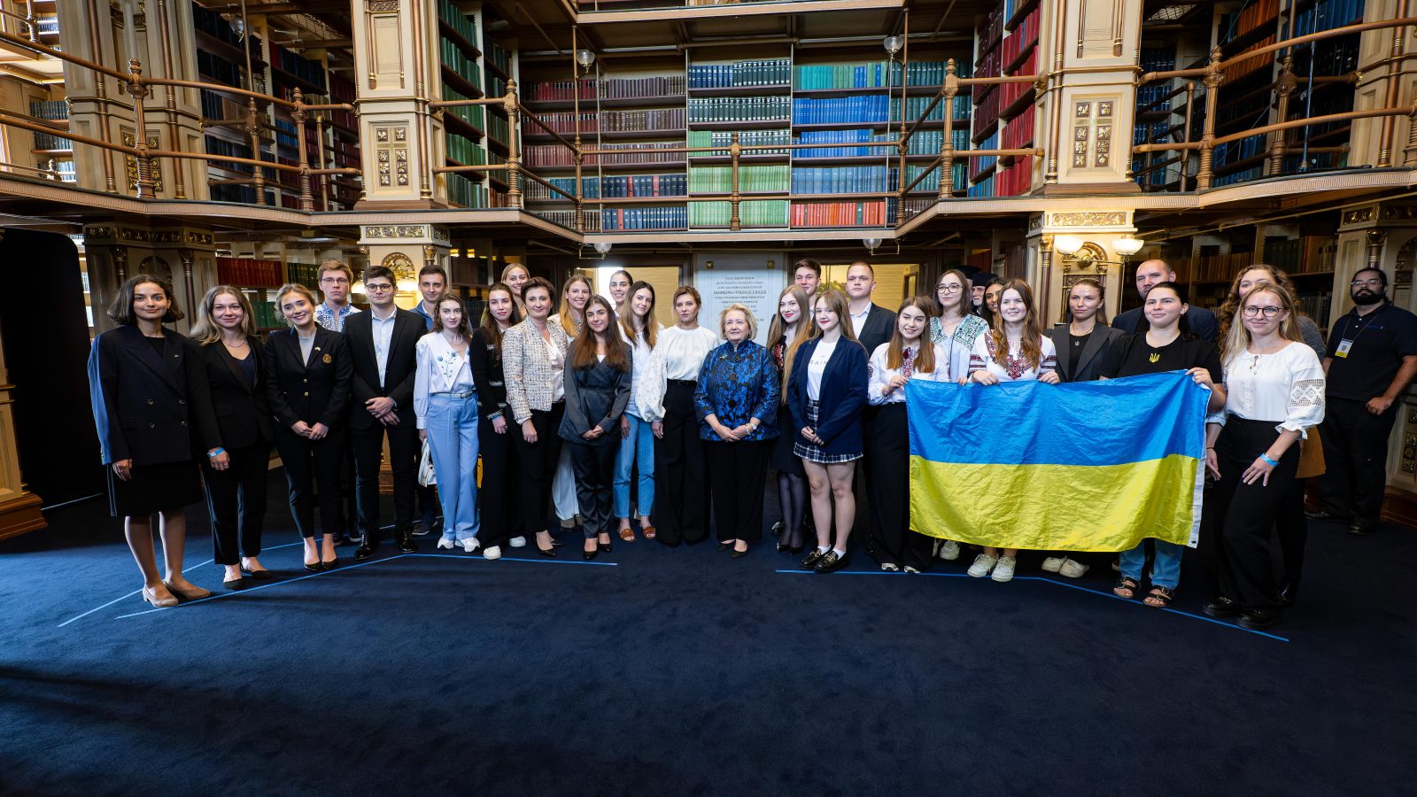 A group of Ukrainian students pose with the First Lady of Ukraine in a Georgetown library. One student holds up the yellow and blue Ukrainian flag.