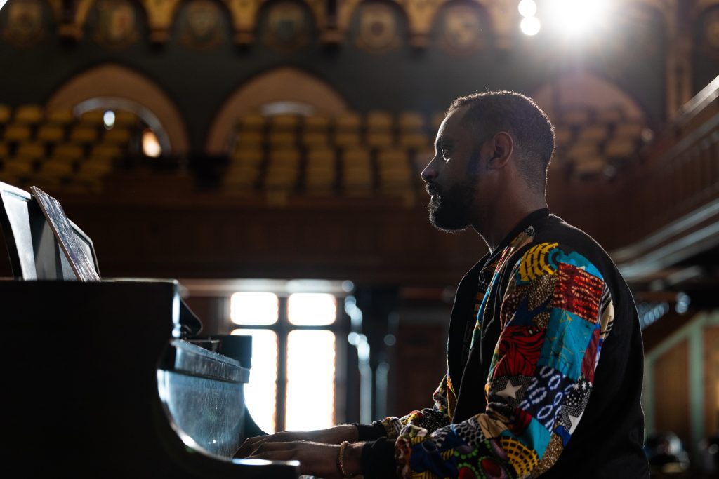 Composer Carlos Simon sits behind a grand piano during a rehearsal in a colorful, patterned jacket with empty seats behind him and sun streaming through the windows of Gaston Hall.