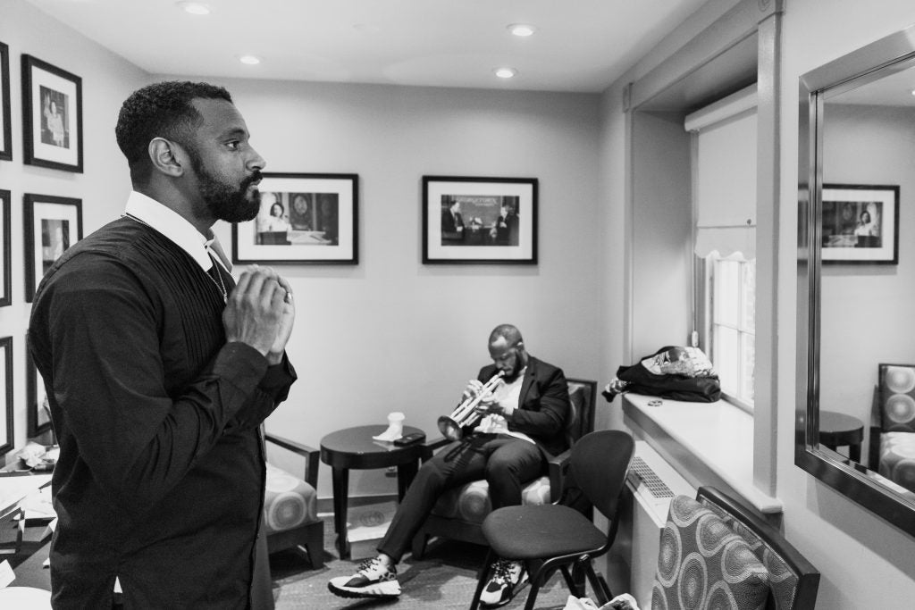 Carlos Simon adjusts his tie while looking into a mirror before his live performance of his requiem in this black-and-white photo. In the corner of the photo, trumpeter MK Zulu practices. 