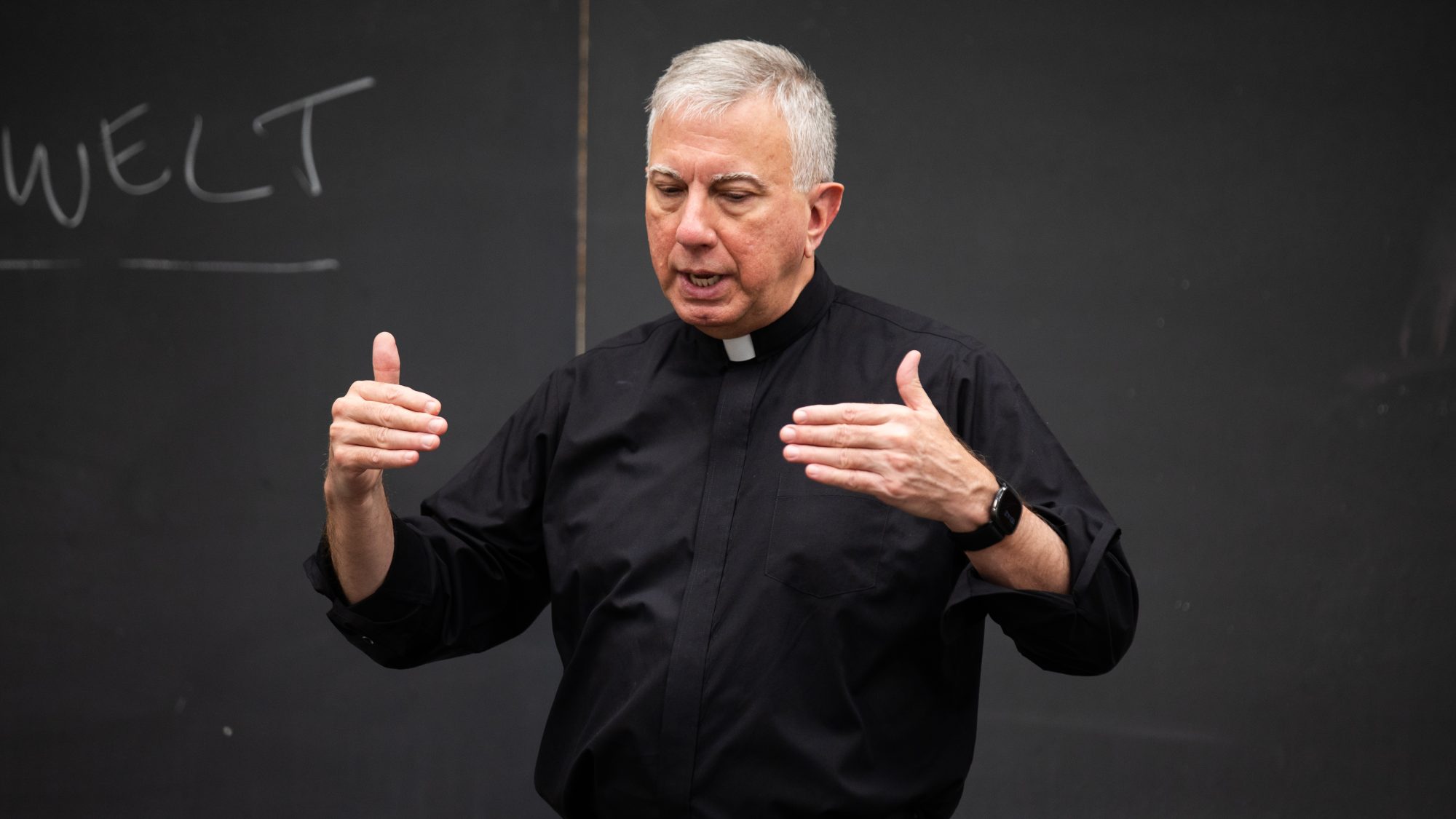 A Catholic priest teaching in front of a chalkboard in a classroom