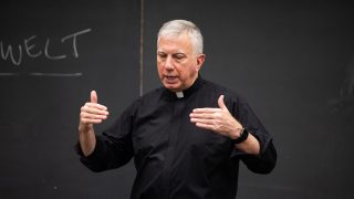 A Catholic priest teaching in front of a chalkboard in a classroom