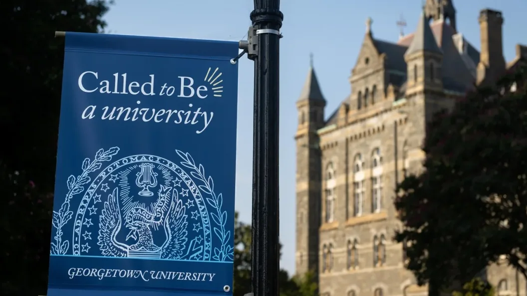 A blue flag hoisted on Georgetown's campus reads "Called to Be a university." Behind the flag is Healy Hall, a historic stone building on Georgetown's main campus.