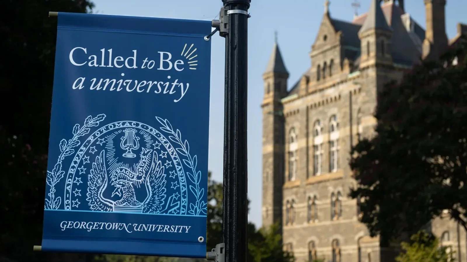 A blue flag hoisted on Georgetown&#039;s campus reads &quot;Called to Be a university.&quot; Behind the flag is Healy Hall, a historic stone building on Georgetown&#039;s main campus.