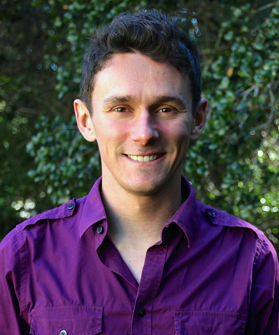 Scot Miller, wearing purple, smiling, standing in front of greenery.