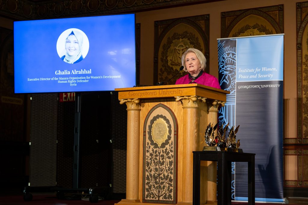 Executive Director Ambassador Melanne Verveer stands behind a podium in Gaston Hall and introduces a video behind her of Ghalia Alrahhal, an award recipient. 