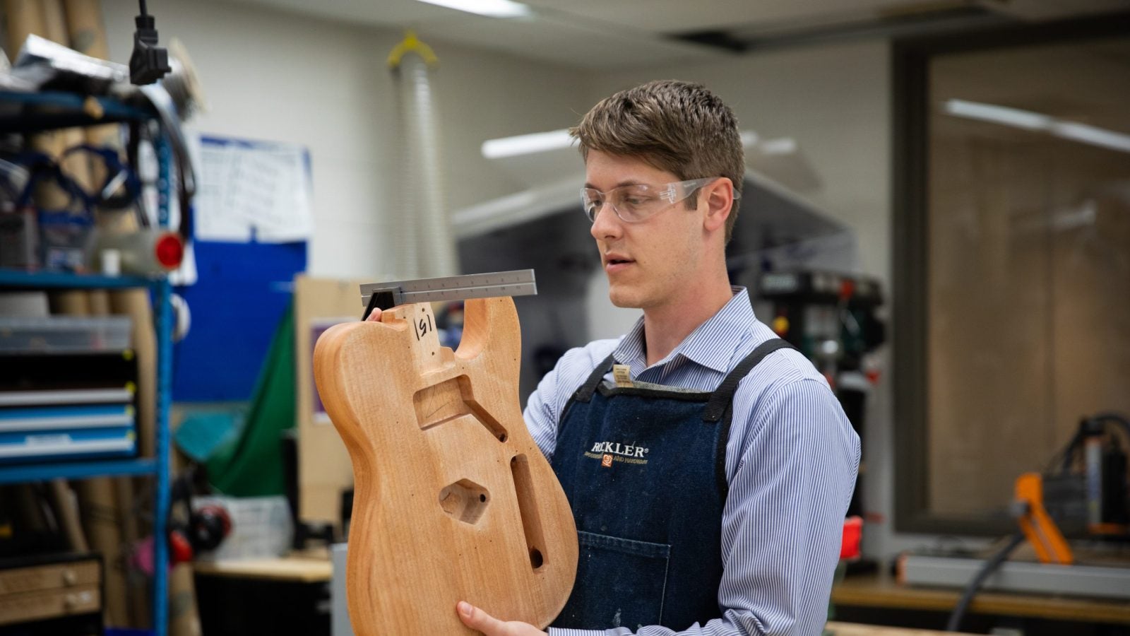 David Strout wears protective glasses and holds up the bottom of a wooden guitar in the Maker Hub on Georgetown's campus.