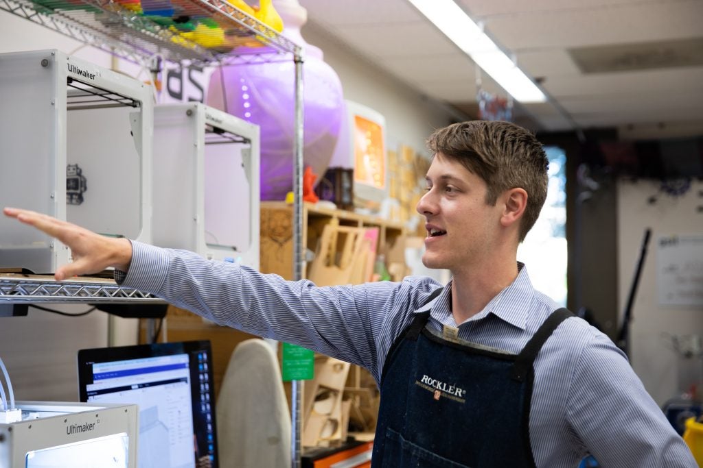 David Strout wears an apron over a collared shirt and points to a 3D printer in the Maker Hub, a collaborative space in Georgetown's Library.