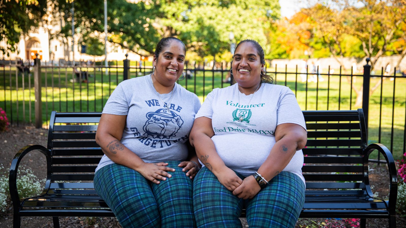 Two sisters wearing matching plaid pants and gray T-shirts sit on a park bench by the front lawn of Georgetown.