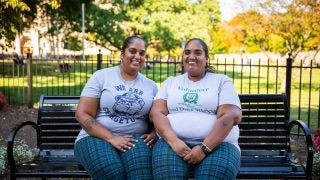 Two sisters wearing matching plaid pants and gray T-shirts sit on a park bench by the front lawn of Georgetown.