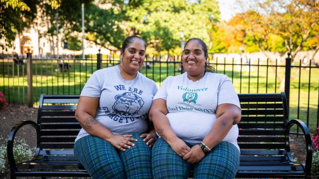 Two sisters wearing matching plaid pants and gray T-shirts sit on a park bench by the front lawn of Georgetown.