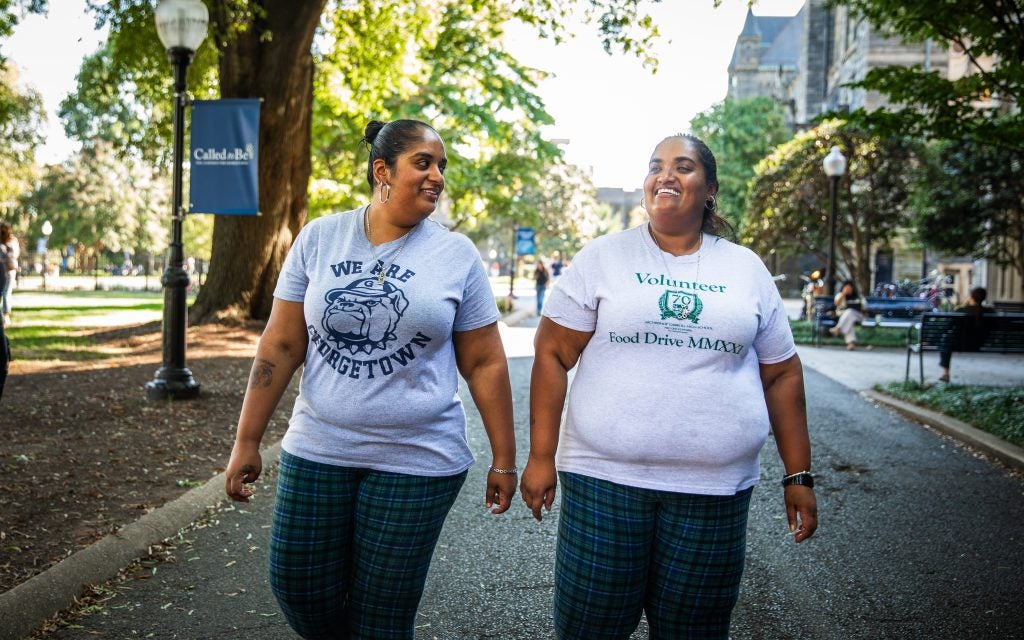 Two sisters walk down a paved walkway on Georgetown's campus. They wear T-shirts and matching plaid pants.