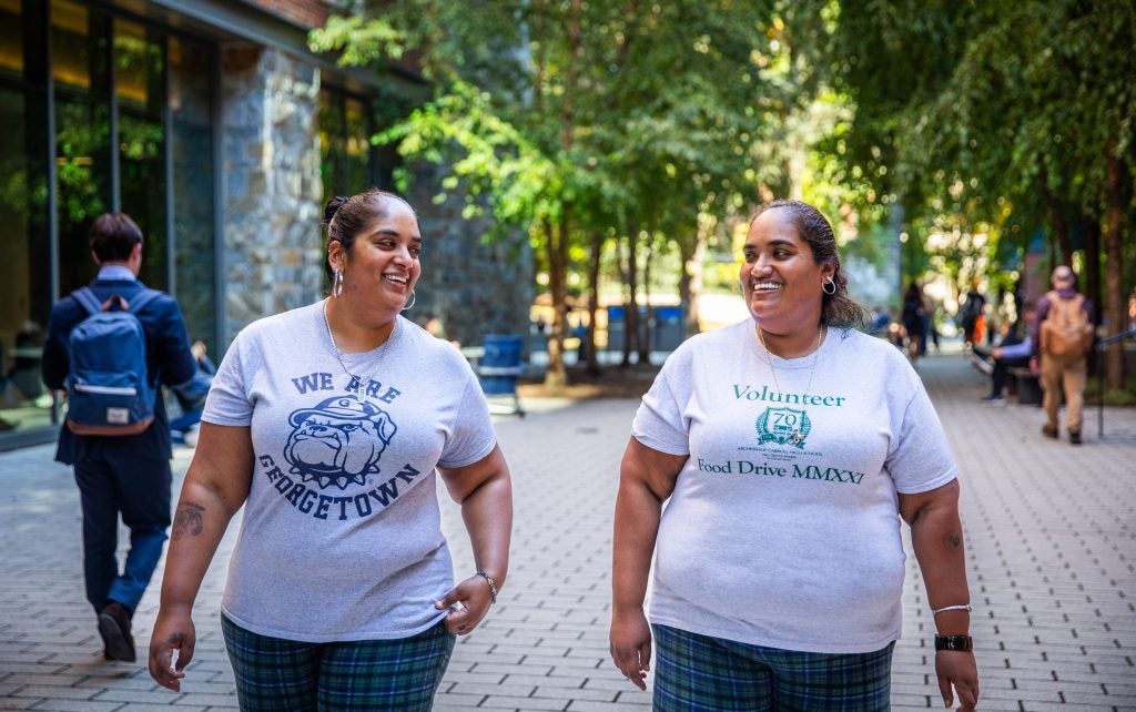 Two sisters smile at each other while walking on Georgetown's campus. They are wearing gray T-shirts and matching plaid pants.