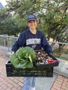 A student wearing a Georgetown baseball gap and sweatshirt holds a tray of fresh vegetables.