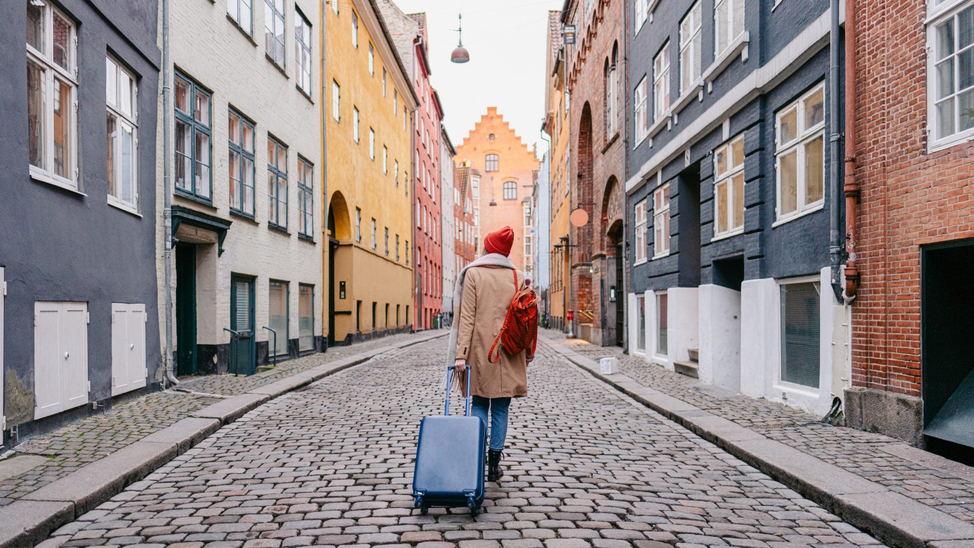 A woman walks down a cobblestone street surrounded by colorful buildings in Denmark. She wears a red winter hat and tows a blue suitcase behind her.