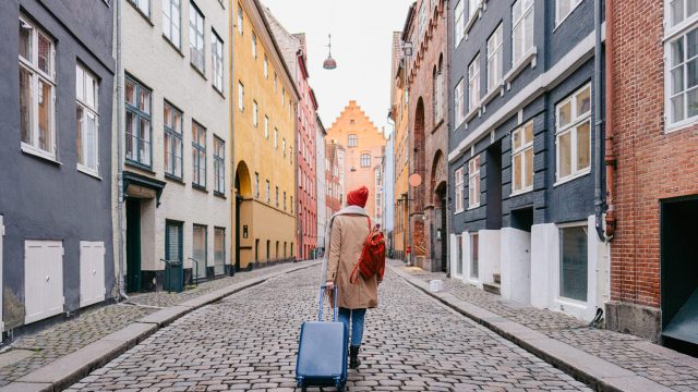 A woman walks down a cobblestone street surrounded by colorful buildings in Denmark. She wears a red winter hat and tows a blue suitcase behind her.