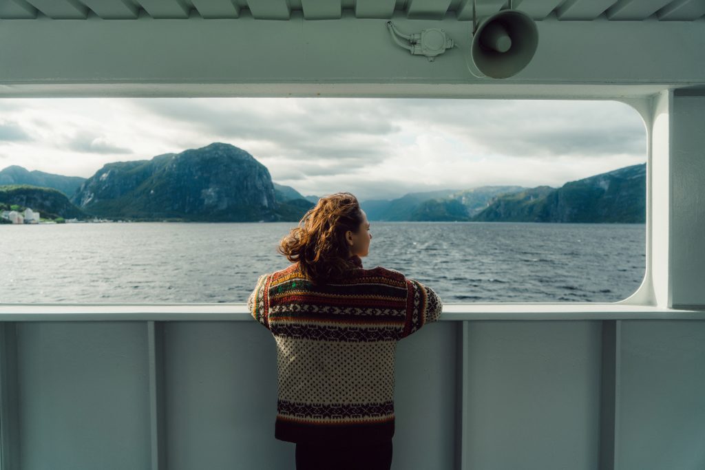 A woman stands with her back to the camera on a ferry and looks out at a view of water, mountains and cloudy gray skies.