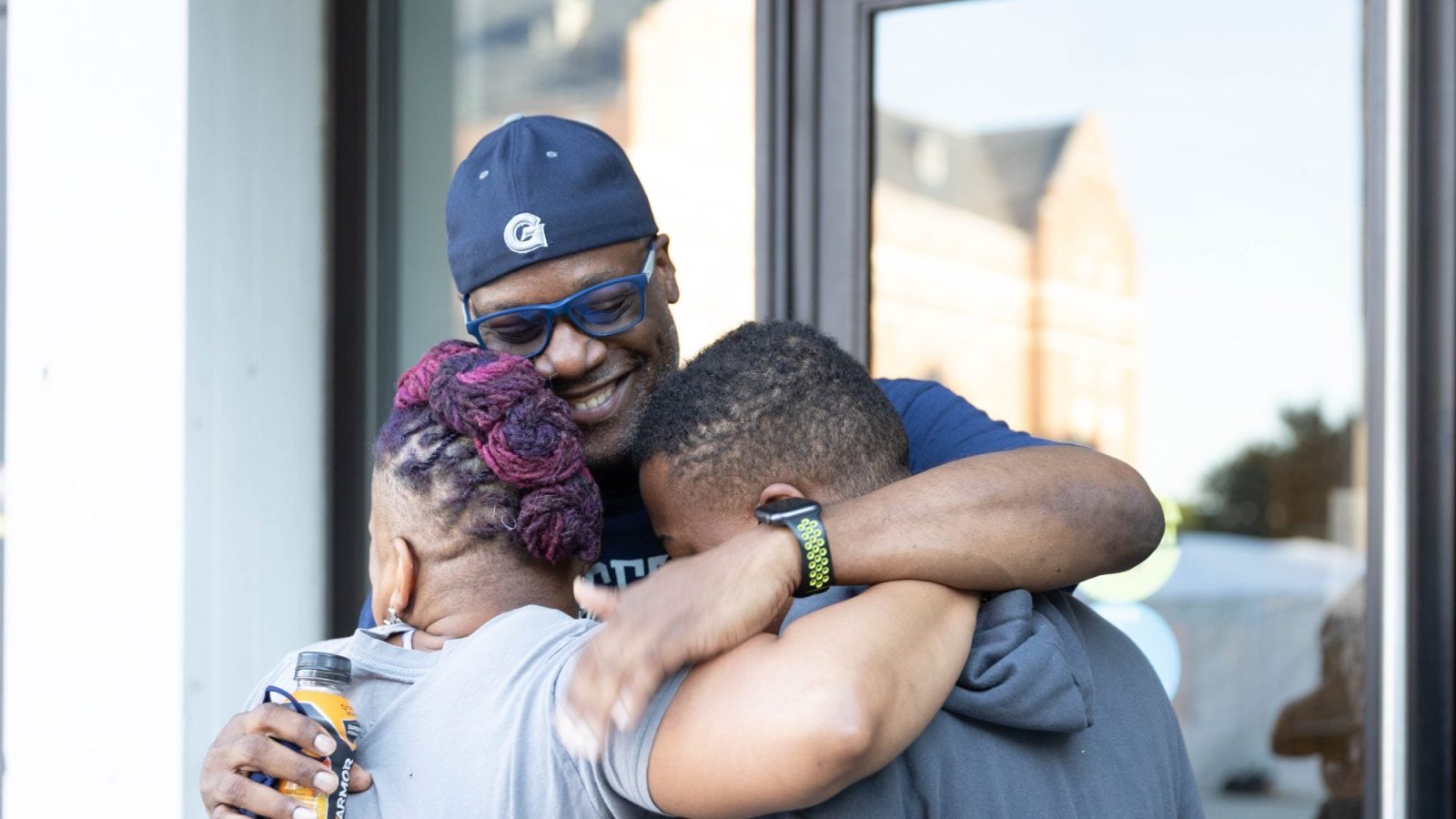 A parent hugs two children on Georgetown&#039;s campus wearing a Georgetown Hoyas baseball cap.