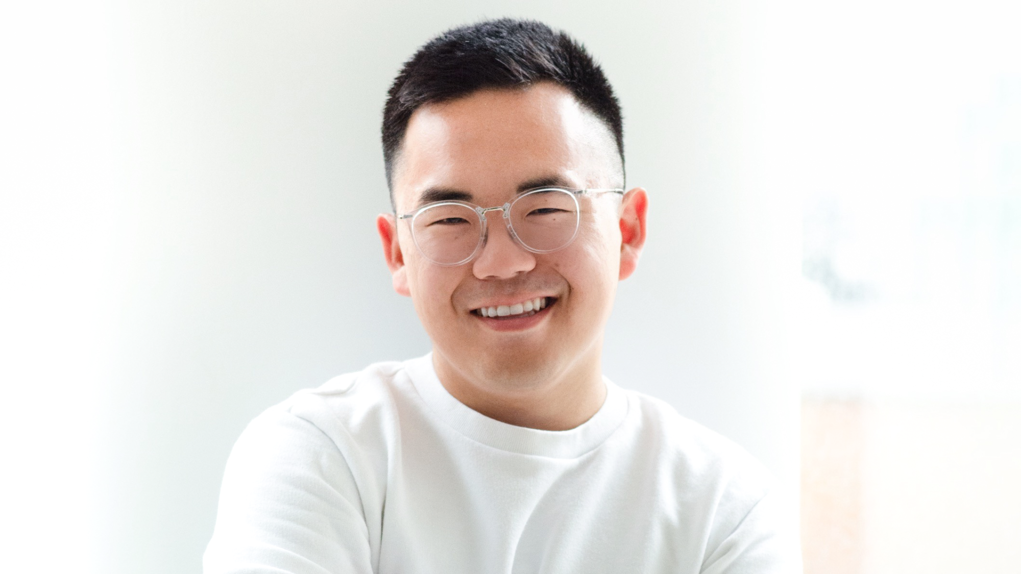 A young man with glasses and short, black hair smiles widely at the camera with folded arms.