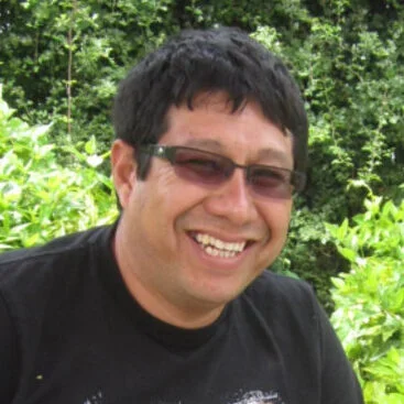 Oswaldo Villena smiling at camera, in front of leafy backdrop.
