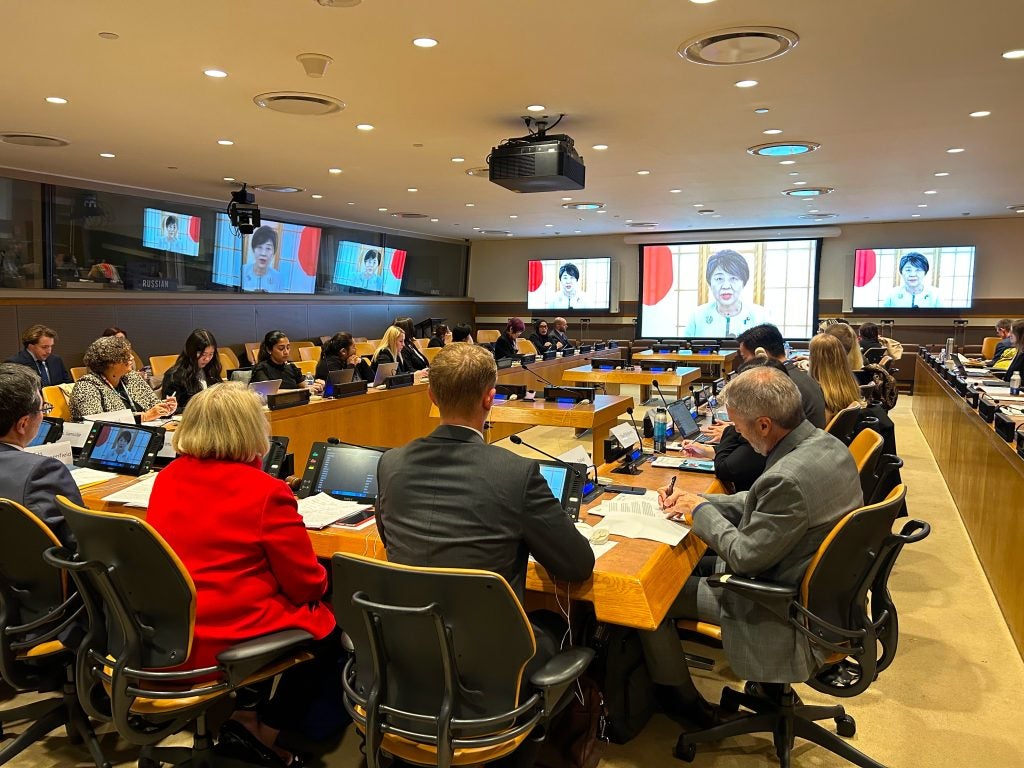 A conference room full of people in a rectangular table facing a tv with a person's face 