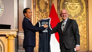 The President of Indonesia, dressed in a black suit, holds up a navy jersey with his last name and number on it. He stands on a stage at Georgetown next to a smiling dean of the School of Foreign Service.