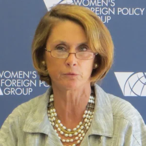 Amb. Barbara Bodine wears a pearl necklace, glasses, and a light green jacket. She answers a question in front of a blue background that says, "Women's Foreign Policy Group."