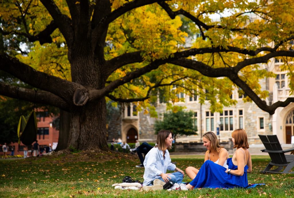 Three white female students sit on a blanket on the front lawn of Georgetown in front of a giant tree.
