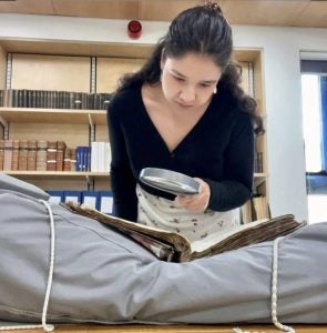 A student uses a magnifying glass to study the contents of an archived record laying on a plump gray cushion in a library.