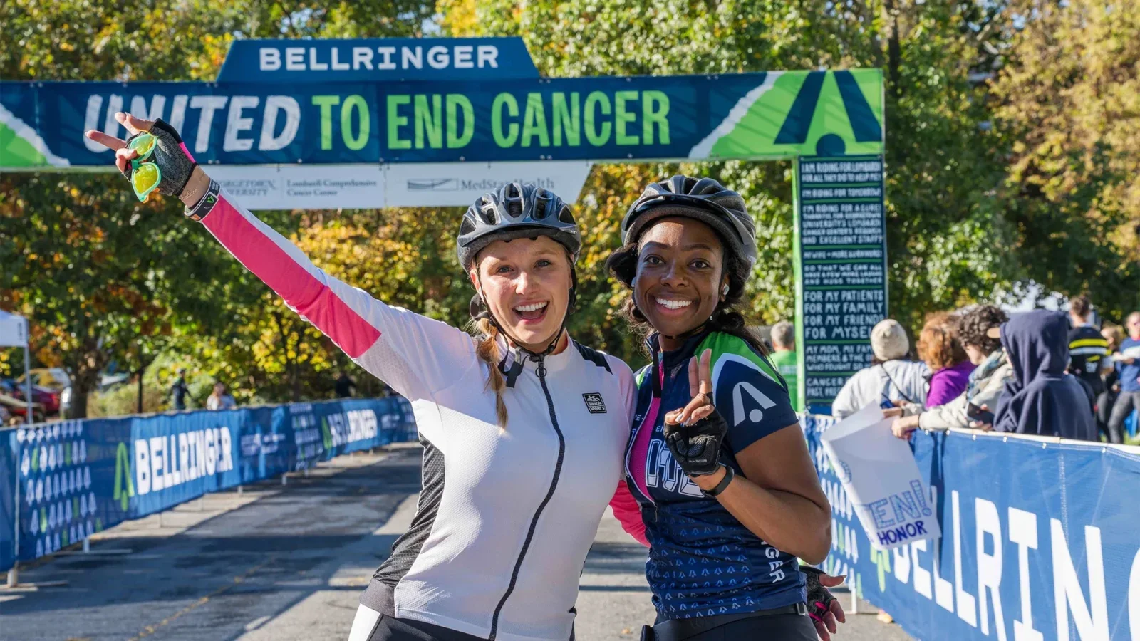 Two bikers wearing helmets and biking gear stand at the finish line of BellRinger 2023, a bike ride to end cancer.