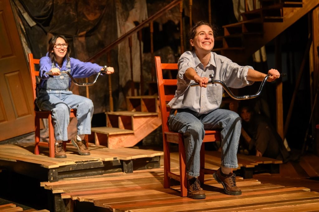 Two students sit on chairs and pretend to ride bikes on stage during a live performance.