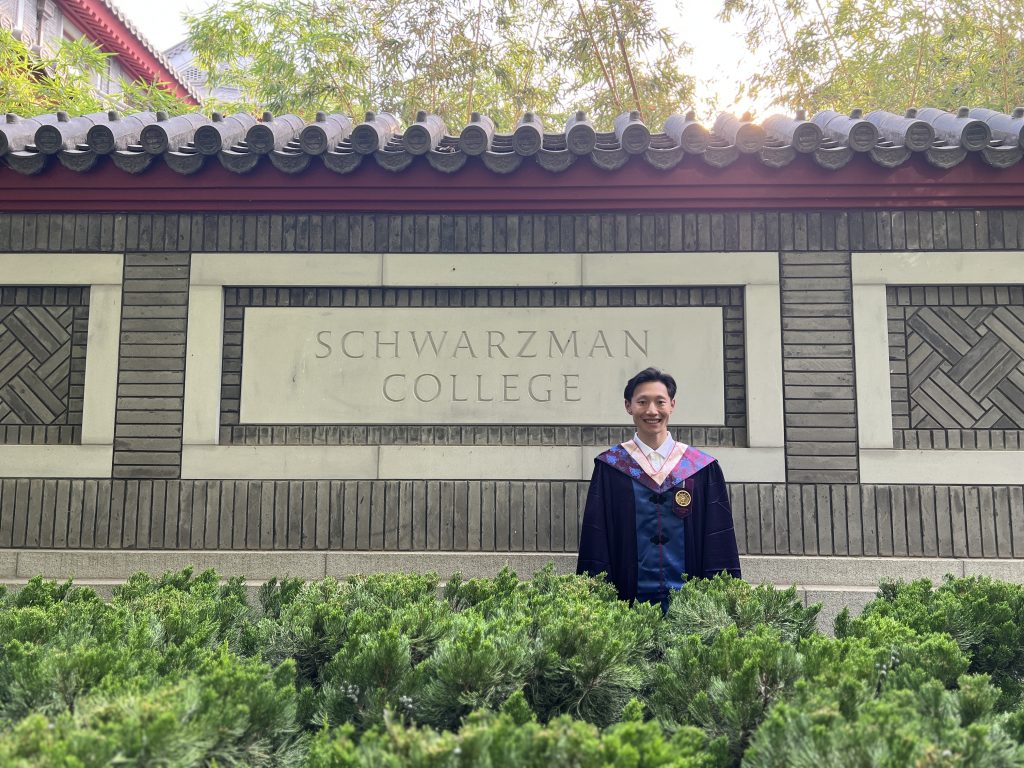 Charlie Wang stands in his graduation robe in front of a stone sign that says "Schwarzman College" in Beijing.