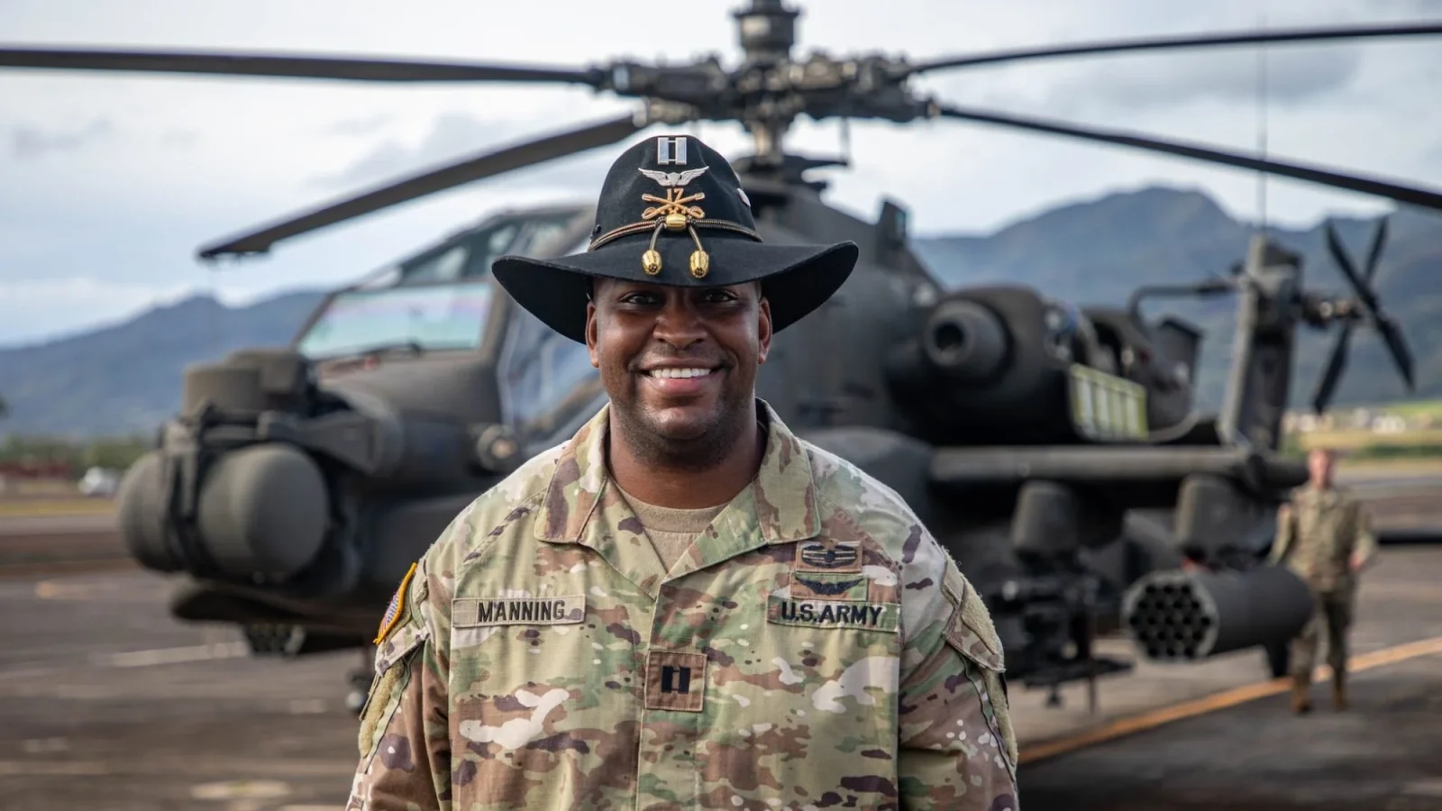 A McCourt Student and U.S. Army Captain stands smiling in front of a helicopter. He is a Black man wearing a hat decorated with military regalia and U.S. Army fatigues.