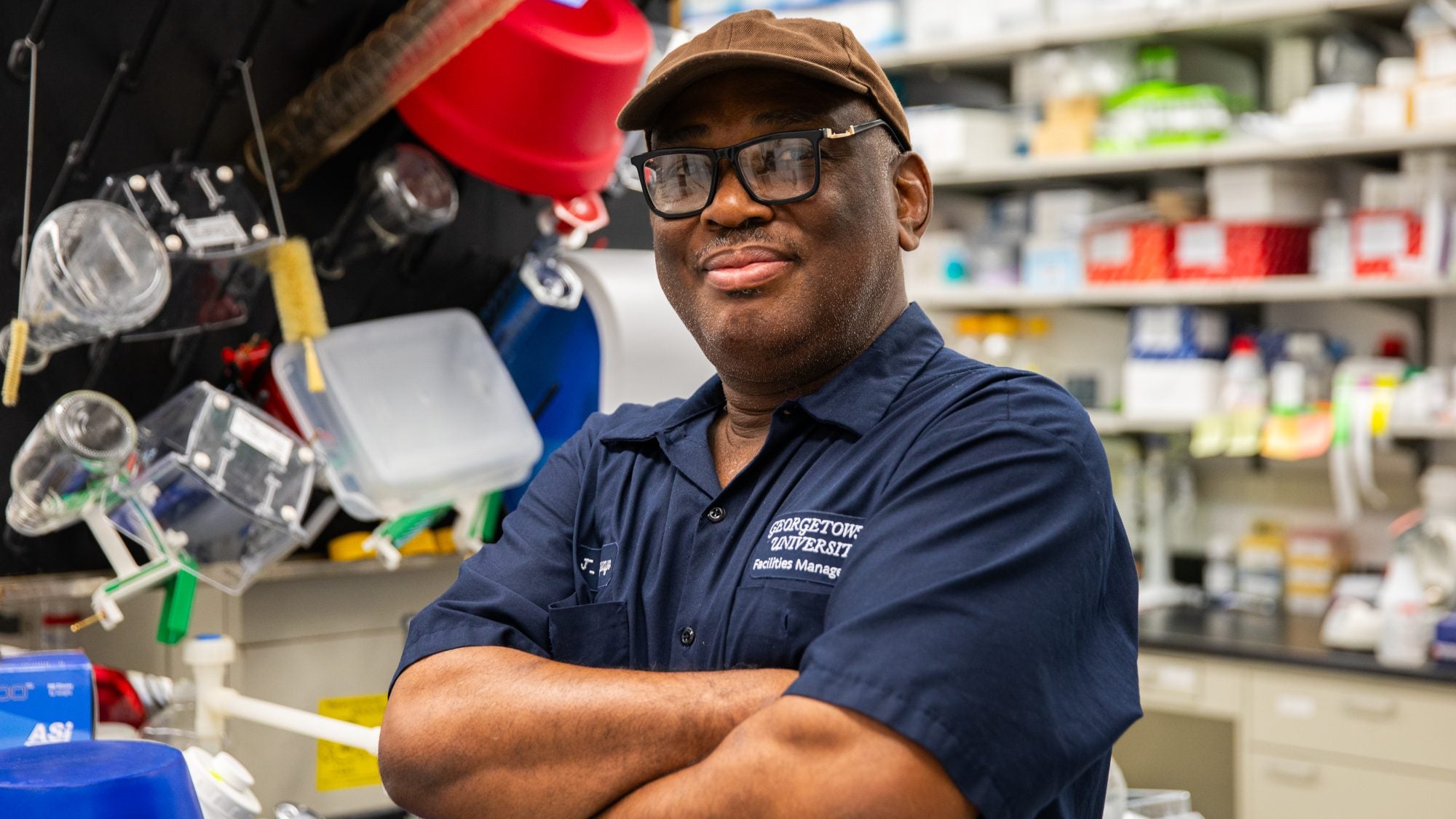 A custodian poses with his arms crossed and smiles slightly at the camera. He wears a navy uniform, a tan baseball cap and black glasses.