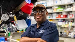 A custodian poses with his arms crossed and smiles slightly at the camera. He wears a navy uniform, a tan baseball cap and black glasses.